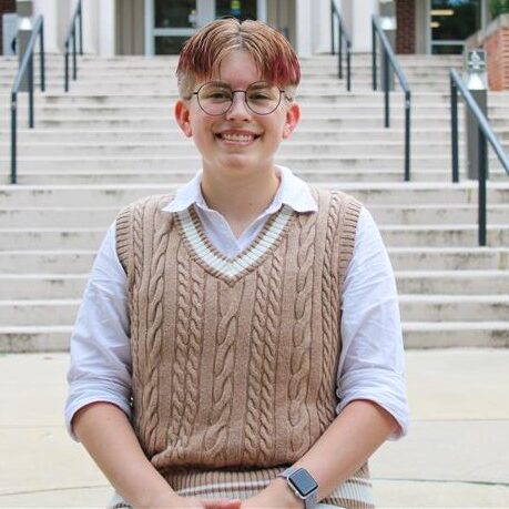 A young man wearing glasses and a sweater vest sits in front of a flight of concrete steps.